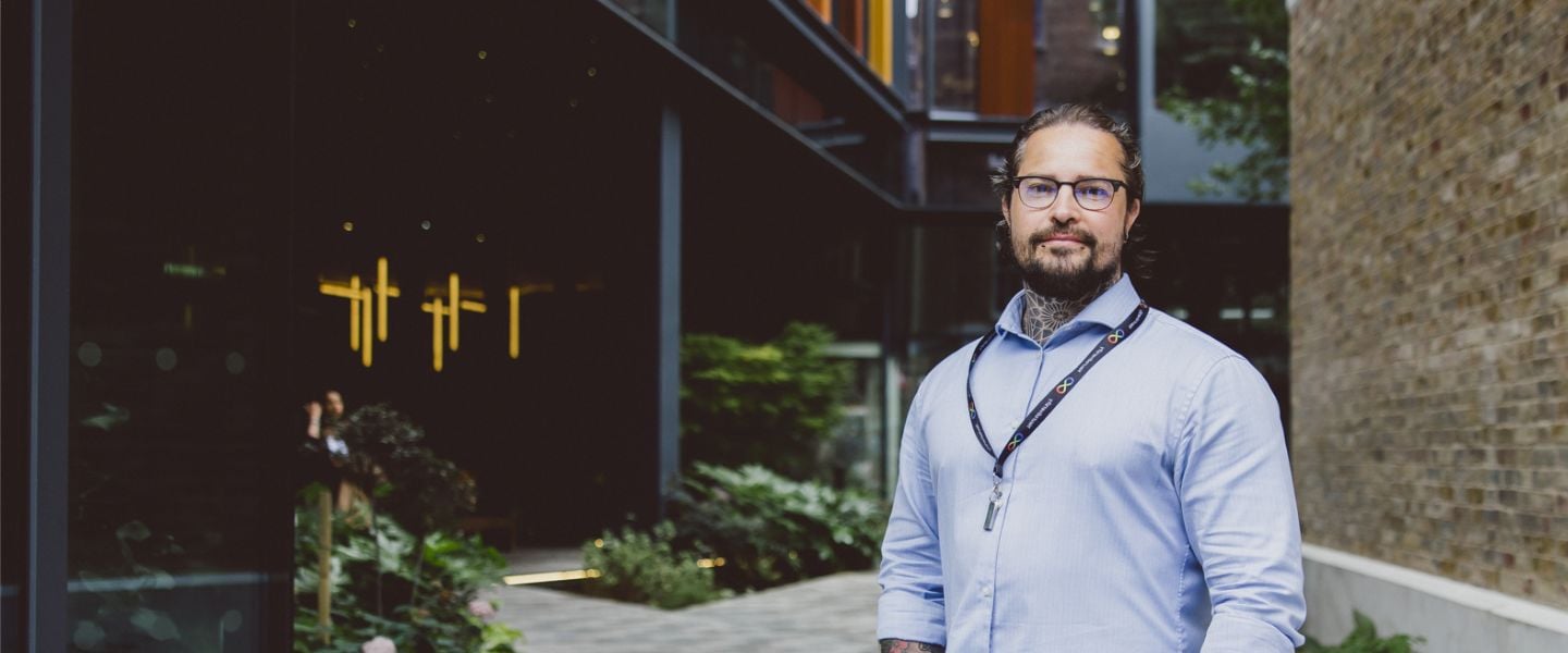 A man is smiling and looking directly at the camera, with his hands tucked into his jean pockets. He is wearing a pale blue shirt, and a lanyard signalling neurodiversity. He has glasses and you can see tattoos at the top of his shirt and on his arms, and two piercings on his chin. He is standing in front of a pale brick wall.