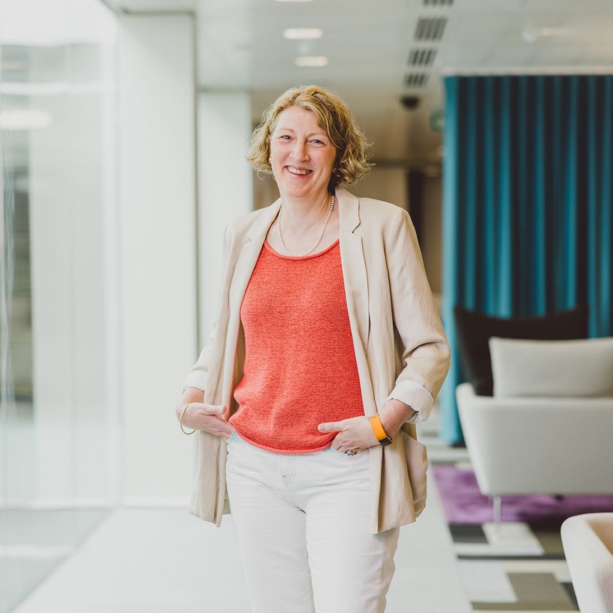 A woman with short cropped hair pictured wearing a coral top and beige jacket, she is in an open office space with glass walls and seating behind her.